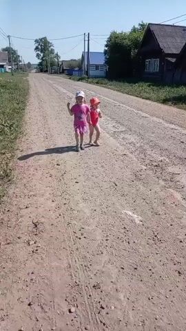 Two young girls walk hand-in-hand down a dirt road. One girl wears a pink t-shirt and shorts with a baseball cap. The other wears a red swimsuit with a matching sun hat. The road is lined with green vegetation on either side and houses are visible in the background. The sky is blue and sunny.