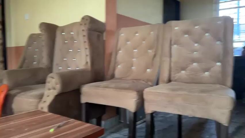 The video shows a living room with brown tufted chairs and a television mounted on the wall. There are framed photos on a black entertainment stand below the TV. The room has a patterned curtain and a door in the background. There is also a small orange chair visible. The floor is tiled.