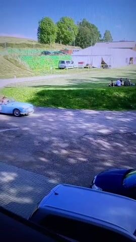 A blue convertible classic car with two occupants drives on a paved road. The car has the number 15 on its side. In the background, there is a large white tent and several people sitting on a grassy hill. Trees and buildings are visible behind the tent. The foreground shows the hoods of two parked cars, one blue and one silver. The sky is bright and clear.