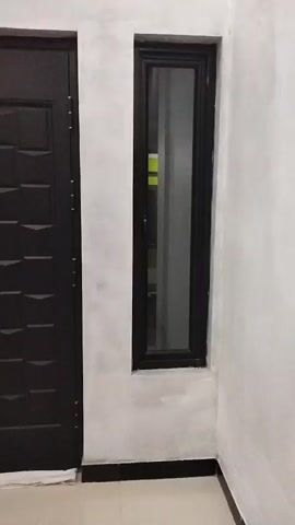 The video shows a close-up view of a dark-colored, paneled door with a metal frame. To the left of the door is a narrow, vertical window with a black frame. The wall surrounding the door and window is painted white, with a rough texture. A small, brown doormat with a decorative border is placed in front of the door. The floor is tiled with a light-colored material. The scene appears to be an interior or exterior entryway. There are no people or animals visible in the video, and no actions or interactions are depicted. The lighting is moderate, and the overall impression is one of a simple, modern architectural design.