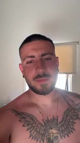 A medium, eye-level shot shows a shirtless man indoors. He faces the camera directly, introduces himself as Radoslav, and says “Dobar den.” He has short, dark hair, a beard, and a mustache. He wears earrings and a gold necklace with a cross pendant. He has a large chest tattoo with wings, a crown, and a skull. Behind him, a window with a partially rolled down shade is visible. A water bottle is visible on a surface behind his left shoulder. The room appears to be lit by natural light.