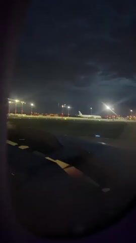 The video provides a night or dusk view from an airplane window, looking out onto an airport tarmac. The wing of the current aircraft is prominently visible in the lower portion of the frame, showing its red navigation lights and parts of its structure.

In the distance, another large passenger airplane can be seen on a runway or taxiway, illuminated by bright floodlights and airport ground lights. The entire airport environment is aglow with numerous red, white, and green runway and taxiway lights, creating distinct lines across the dark landscape. The sky above is dark and cloudy, with hints of an overcast atmosphere.

The primary activity depicted is the plane from which the video is taken taxiing on the airport grounds, likely either before takeoff or after landing. The audio features a distinct "woo" sound, indicating excitement or joy from the person recording the video, possibly related to travel or the anticipation of a flight. No visible text appears in the video itself.

The scene is set in an airport environment near Ecser, Hungary, strongly suggesting Budapest Ferenc Liszt International Airport (BUD).