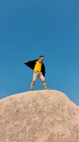 The video features an adult male standing atop a rounded, light-colored rocky hill under a vast, clear blue sky. He is dressed in a yellow t-shirt, a dark jacket, khaki pants, and brown shoes.

The activity showcases a sequence of dynamic poses performed by the man. Initially, he stands angled, looking to his right with a dramatic posture. He then transitions into a pose facing the camera, with arms spread wide and mouth open, suggesting a shout or roar. Subsequently, through a quick edit, he is seen holding a large, dark, rectangular object (possibly a jacket or blanket) over his head and back, conveying an impression of lifting or carrying something heavy, while maintaining an intense expression.

Notable details include the bright, clear daytime weather with no clouds visible. The rocky, conical formation hints at a distinctive natural landscape, consistent with the geological features found in Ürgüp, Turkey. The man's expressions throughout the video are energetic and dramatic, suggesting a performance or staged moment. There is no visible text in the video.