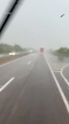 A windshield view of a highway during a rainstorm. Water droplets streak down the glass, obscuring visibility. A red truck is visible in the distance, its taillights illuminated. The road is wet and reflective, with white lane markings visible. Trees and foliage line the sides of the highway. A windshield wiper sweeps across the frame in the foreground. The sky is overcast and gray.