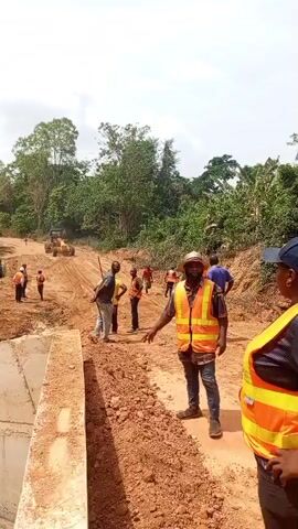 The video captures various scenes from an outdoor road construction site in Aloko, Nigeria, during daylight hours. The ground is predominantly reddish-brown earth, with lush green vegetation, including numerous palm trees and banana plants, bordering the site.

The initial scene shows a group of individuals, primarily men and one woman, at the construction site. Several people are wearing bright orange safety vests, suggesting a work or supervisory role. In the foreground, a man in a safety vest and hat is seen conversing with a woman also in a safety vest and a blue cap. Other workers are in the background, near what appears to be a large construction vehicle, possibly a bulldozer or grader. A concrete structure, possibly part of a culvert or bridge abutment, is visible in the lower left. The woman's black shirt features white text, appearing to be "ARMANI."

Subsequent clips provide different perspectives of the setting. Two men, one shirtless and the other in a dark t-shirt, are observed standing and talking on a cleared patch of dirt surrounded by dense tropical foliage. A wider shot showcases the verdant environment, dominated by palm trees and other plants.

The final segment shows two men standing on the unpaved road. One, wearing a white tank top, looks towards the camera. The other, in a brown t-shirt, holds a long stick or tool, possibly a rifle. In the background, a white pickup truck and an orange dump truck are visible on the dusty road, suggesting ongoing construction activity. The weather appears sunny or partly cloudy, indicating clear working conditions. The overall impression is that of active road development in a rural, tropical setting.