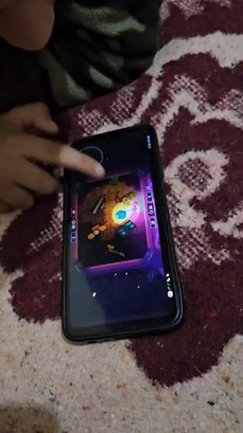 A child is playing a mobile game on a smartphone while lying on a bed. The phone displays a top-down view of a game that appears to be a puzzle or strategy game with various objects arranged within a square border. The child's finger is touching the screen, presumably interacting with the game. The bedspread has a maroon and cream-colored pattern. The child is wearing a green and brown camouflage patterned garment. The room is lit well enough to show the details of the phone and the child's hand.
