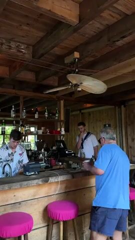 The video shows an interior scene of a bar with a rustic wooden ceiling and walls. A ceiling fan with wooden blades is visible in the center. In the foreground, there is a wooden bar with three pink stools. A man in a blue t-shirt and shorts is seated on one of the stools, facing away from the camera. He is wearing sunglasses. Behind the bar, a younger man in a white shirt with a blue collar and a white sailor-style hat is standing. Another man, possibly a bartender, wearing a patterned shirt is also behind the bar, to the left. There are shelves with bottles of alcohol and coffee-making equipment behind the bar. The man in the blue t-shirt appears to be looking at something on the bar or at the bartender. The lighting is natural, suggesting it's daytime.