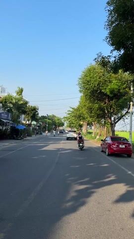 The video shows a street in what appears to be a suburban or rural area. The road is gray asphalt, with double yellow lines down the center. There are people driving motorbikes, a bicycle, and a black SUV. Trees line one side of the street. On the other side, there are businesses and houses. The sky is blue. A red car is parked on the side of the street. A rice field is adjacent to the trees on the right side of the street. The wind appears to be blowing strongly, creating some audio distortion.