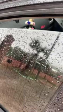 The camera is positioned outside a parked car during rainy weather. The vehicle's window is slightly ajar, allowing a partial view of the interior. The interior features black upholstery, a steering wheel with the Volkswagen logo, a dashboard with the word "GOLF" in red lettering, and a central display screen. Raindrops are visible on the window, obscuring the view of the exterior, where a brick building and some trees can be partially seen. The scene appears to be a still shot, with no visible movement or activity other than the rain.