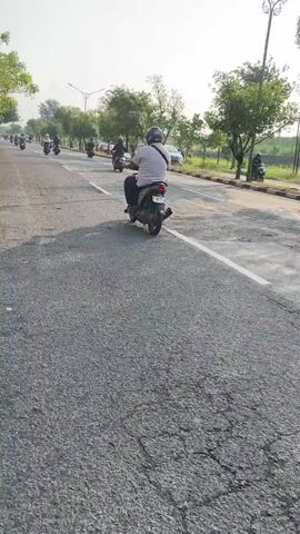 A group of people are riding motorcycles on a paved road. The road has some cracks and potholes. There are trees and grass on the side of the road. The sky is clear and the sun is shining.