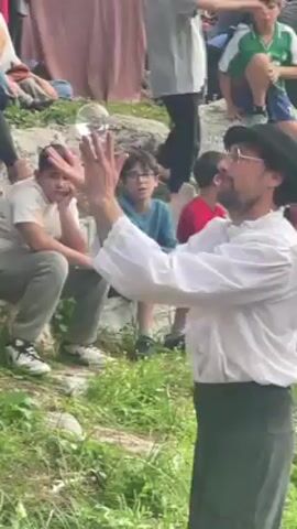 The video shows a performance at an outdoor fair, with an adult male performing a magic trick involving a large, clear ball. He wears a white, frilly shirt, black pants, and a black hat with red accents. The performer manipulates the ball between his hands, seemingly making it float. In front of him, a small group of young children are sitting on rocks. One child looks bored or disinterested, resting his head on his hand. Another child wears glasses and watches with a more interested expression. There are other people milling around in the background, including some looking at merchandise displayed on tables. The environment appears to be grassy and somewhat uneven.