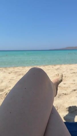 The media file shows a first-person perspective of someone relaxing on a sandy beach. The foreground is dominated by the person's legs and knees, with sand clinging to their feet. In the background, a clear, turquoise ocean stretches out to the horizon, meeting a bright blue sky. The sun is shining, indicating a warm, clear day. Several people are visible in the shallow water, suggesting it is a public beach. The overall mood is one of relaxation and enjoyment of a beach vacation. The scene is set on a beach with fine sand and calm, shallow water. There is no visible text in the media file.