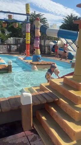 A child wearing a colorful rash guard and a blue hat is crawling on the edge of a shallow swimming pool, reaching for a yellow object. The pool is part of a water park with water slides and totems with faces in the background. Several other people are visible in the water and near the slides. The sky is blue with scattered clouds, and trees are visible behind the water park. The scene is brightly lit by sunlight.