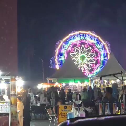 The video shows a night scene at an outdoor market or fairground. A large Ferris wheel, illuminated with colorful lights, is visible in the background. In the foreground, there are numerous stalls set up under tents, displaying various goods. People are seen walking around and browsing the stalls. The atmosphere appears lively and festive.