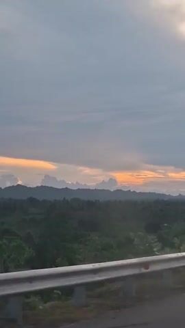 The video shows a view from a moving vehicle on a road during a cloudy sunset. A guardrail is visible in the foreground, separating the road from a landscape of trees and rolling hills. The sky is filled with gray and orange clouds, indicating the sun is setting. There are no people or animals visible in the scene. The camera appears to be pointed towards the horizon, capturing the expansive view of the natural landscape and the sky.