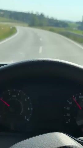 The video shows a first-person view from inside a car as it drives along a winding road. The steering wheel and dashboard are visible in the foreground. The speedometer shows the car is traveling at approximately 80 km/h. The road is paved with white lines and curves to the left. Outside the car, there are green fields, trees, and a clear blue sky. The sun is shining, indicating it is daytime. There are no other people or vehicles clearly visible in the immediate vicinity. The scene suggests a typical drive through a rural or semi-rural area.