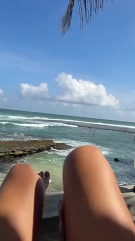 The video shows a first-person perspective of someone relaxing by the ocean. The camera focuses on their legs and feet, which are tanned and appear to be sun-kissed. The legs are positioned at an angle, with one leg extended and the other bent. In the background, the ocean is visible with waves gently crashing onto a rocky shore. The sky is a clear blue with a few white clouds. A palm frond is partially visible at the top of the frame, suggesting a tropical location. The overall impression is one of relaxation and enjoyment of a seaside environment.