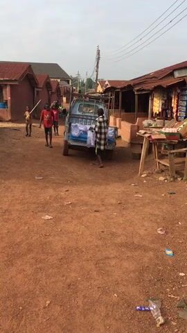 The video shows a dirt road in what appears to be a village. A small blue truck is parked in the middle of the road with several bags of water bottles in its bed. A man in a plaid shirt is carrying one of the bags away from the truck. Several children walk around the area, including one carrying a long stick. There are small buildings on either side of the road, some with tin roofs. Debris litters the ground.