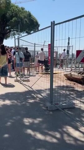 The video shows a group of people walking along a paved path towards a barrier. The barrier appears to be a metal fence section, possibly indicating a restricted area. There are trees casting shadows on the path. The people are casually dressed in summer clothing. Some appear to be queuing near the barrier. There's a water fountain beside the path. The weather looks sunny.