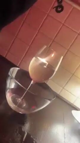 A hand holds a pair of metal tongs above an upside-down wine glass containing a tea light, submerged in water. The wine glass rests on the rim of a glass bowl. The scene is lit with a warm, reddish light, casting shadows on the tiled wall and dark countertop.