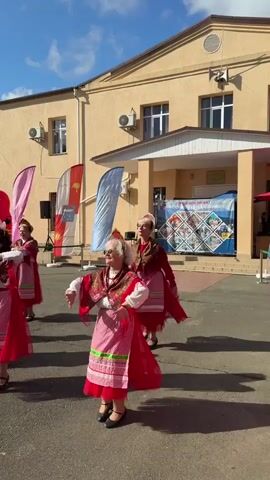 Five elderly women in traditional red and white Russian folk costumes are dancing outdoors in front of a yellow building. The women are all wearing red dresses with white blouses and red aprons with white and green trim. They have their arms raised and are smiling. In the background, there are flags and banners, and the sky is blue with some clouds. The ground is asphalt.