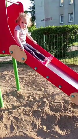 A young girl with blonde hair, wearing a grey t-shirt, black leggings with white polka dots, and pink shoes, is on a red playground slide. She is sitting at the top of the slide, holding onto the sides, and appears to be looking towards the camera. The slide is on a sandy playground, with a green fence and a building in the background. The girl then slides down the slide and stands up at the bottom, facing the camera. She then puts her hands on her hips and turns to the side. The sun is shining brightly, creating shadows on the sand.