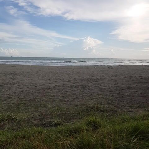 The video shows a beach scene. On the left side of the frame, there is a sky with clouds and the sun is visible. The ocean is in the middle with waves. The right side of the frame shows a sandy beach and green grass. The video appears to be taken from a stationary position. There are no people or distinct objects visible in the scene, other than natural elements.