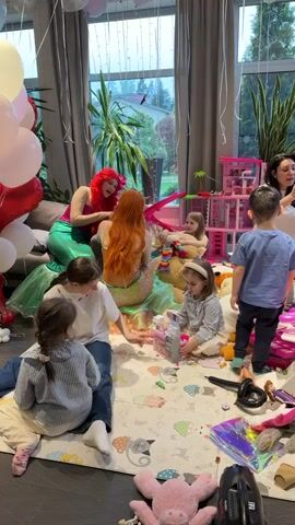 The scene captures a festive indoor gathering, likely a children's party. Several children are playing on a patterned playmat in the foreground, surrounded by toys. Two adults dressed as mermaids, one with red hair and the other with an orange wig, are interacting with the children. One child is standing and looking at the scene, while others are sitting on the playmat engaging with toys. An adult woman stands in the background, partially visible. The room is decorated with balloons, and large windows provide a view of an outdoor area with greenery. The atmosphere seems joyful and playful.