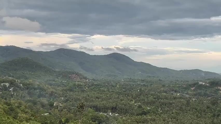 The video presents a wide, static shot of a lush, hilly landscape under a cloudy sky. The hills are covered in dense green vegetation, likely trees and other foliage. Amidst the greenery, there are sparse signs of human habitation, with a few scattered buildings visible. The sky is overcast with dark gray clouds, although a small patch of lighter sky is visible in the distance. The scene lacks any human presence or activity. The overall impression is one of a serene, natural environment, possibly a tropical or subtropical region.