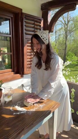 A woman in a white dress and headscarf is kneading dough on a wooden table dusted with flour, outdoors on a porch. A pitcher with lemon water and a plant are on the table. She looks down at the dough as she kneads. The background shows a window with wooden shutters and greenery.