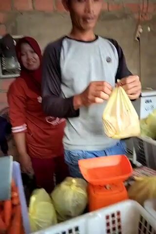 A man in a gray and black long-sleeved shirt and blue jeans is holding a yellow plastic bag filled with produce. He is standing in front of a brick wall and a counter displaying various fruits and vegetables, including carrots, papayas, and what appears to be corn. To his left, a woman wearing a red hijab and an orange top with white stripes is smiling and looking at the camera. She is standing behind a counter as well. An orange weighing scale is visible on the counter in front of the man, along with more produce in plastic bags and white baskets. In the background, there is a white refrigerator with a GE logo, a purple fan, and some shelves. The scene appears to be an indoor market or a small store.
