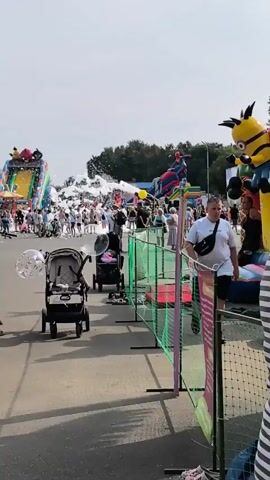 The video shows an outdoor event with several inflatable attractions, including a large slide with a Spider-Man figure on top. There are many people in attendance, including families with children and strollers. A foam machine is visible, producing a large cloud of white foam that is dispersing into the air. In the foreground, two strollers are parked, and a person's arm and shoulder are visible on the right side of the frame. The environment appears to be a paved area with trees in the background, suggesting a park or fairground setting. The lighting indicates it is daytime.