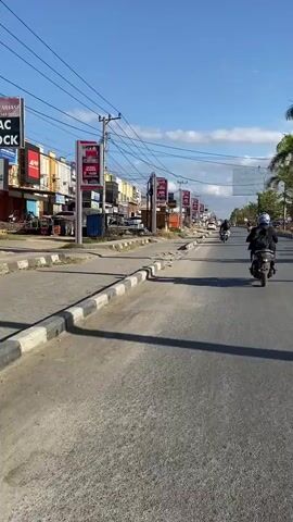 A motorcycle with a rider wearing a helmet and backpack is traveling on a paved road. The rider is in the right lane, and the motorcycle is moving away from the camera. The road is lined with buildings and shops on the left side, with a sidewalk and utility poles. There are other motorcycles and vehicles in the distance. The sky is blue with some clouds.