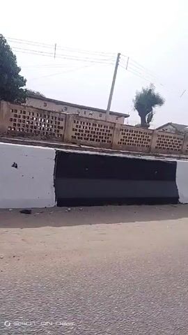 The video shows a bright yellow, three-wheeled motor vehicle, commonly known as a "keke" or "tuk-tuk", driving on a road with a white and black concrete barrier on one side. The barrier is damaged in several places. Behind the barrier is a tan building with a patterned brick wall and a tree. The scene appears to be an urban or suburban setting under a bright, slightly hazy sky.  The keke has the number "901" printed on the front. There is trash and debris on the ground near the barrier. The video is taken from a moving vehicle.