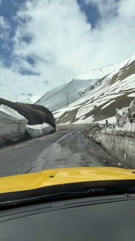 The video shows a view from inside a yellow car driving on a mountain road. The road is paved but has many potholes and patches of snow and debris. On either side of the road are large piles of snow and concrete barriers. In the background, a snow-covered mountain rises to the sky, which is blue with scattered clouds. A few power lines are visible against the mountain. The car seems to be driving slowly and carefully.