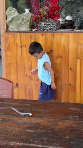 A young boy, wearing a light blue sleeveless top with "AOG" faintly visible text and dark blue shorts, stands in an indoor or semi-covered area. He is leaning against a vertical wooden paneled wall or counter and looking downwards. The setting appears to be a rustic space, possibly a restaurant or guesthouse, with prominent wooden architecture.

Behind the boy, on a ledge above the wooden paneling, are decorative items including red berries, golden dried leaves, a white vase, and a large rock. To the left foreground, a brown plastic chair is visible. In the immediate foreground, a dark wooden table features a silver spoon resting on its surface. The lighting suggests it is daytime. The boy remains relatively still, with a neutral expression, making only a slight movement with his right hand during the video. The environment aligns with the provided context of Du Già Commune, Vietnam.