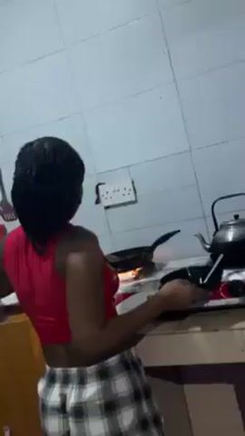 A woman with braids wearing a red tank top and plaid pants is standing in a kitchen. She is facing away from the camera and toward a stovetop with a gas burner lit beneath a black frying pan. She is using a spoon to stir the contents of the pan. To the left of the pan is another frying pan. A white electrical outlet is visible on the wall behind the stovetop. The walls are tiled.