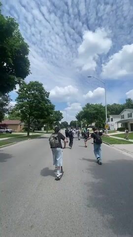The video shows a group of people riding on scooters down a suburban street on a cloudy day. The camera is positioned behind the people, giving a view of their backs as they move away from the camera. The street is paved and lined with houses and trees. Some people are wearing backpacks, and others are wearing casual clothing. The sun is visible through the clouds, casting shadows on the street. The overall scene suggests a leisurely activity in a residential area.
