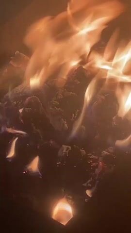 The video shows a close-up view of a fire burning. The flames are a mix of orange and yellow, and they dance and flicker over a pile of what appears to be burning logs or coals. The background is dark, suggesting the fire is in a dimly lit area, possibly outdoors at night or in a fireplace. There are no people or other objects clearly visible in the frame. The focus is entirely on the natural phenomenon of fire.