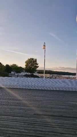 The video pans across a wooden pier or boardwalk with a beach and ocean in the background. Two large white flags on tall poles are visible, one on the left and one on the right. In the middle distance, there is a white structure that appears to be a seating area or a decorative element. Several people are scattered along the pier and on the beach. There are also street lamps visible along the pier. The sky is blue with some scattered clouds, and the sun appears to be low in the sky, suggesting it is either early morning or late afternoon. The overall scene depicts a public waterfront area during a pleasant time of day.