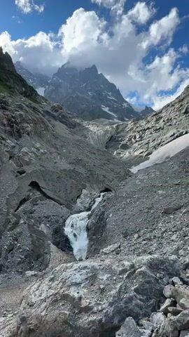 The video shows a view of a mountain landscape. In the foreground, there are large rocks and gravel. A stream of white water flows down the rocks in the middle of the frame. In the background, a large, imposing mountain range rises up, with its peaks partially obscured by clouds. The sky above is blue with scattered white clouds. The scene appears to be a natural, rugged environment, likely a mountainous region.