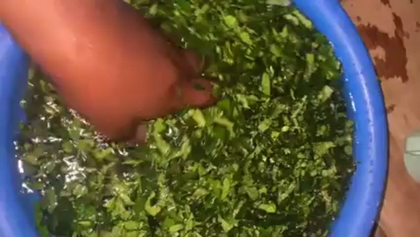 A top-down, close-up shot shows a dark-skinned hand in a blue plastic basin filled with water and chopped green leafy vegetables. The hand is moving, appearing to wash or agitate the vegetables in the water. Some leaves are held between the fingers for a moment.  A small white object is visible at the edge of the basin, possibly a container or utensil. The basin sits on a surface with a slightly mottled brown pattern.