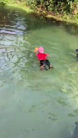 A child wearing a red shirt, black shorts, black shoes, and a pink swim cap swims in a green-tinted body of water. The child is holding a yellow object above the water with one arm. The body of water is surrounded by lush green vegetation. The child moves through the water using a paddling motion with their arms and legs.