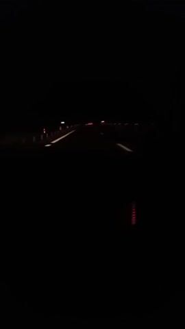 The video shows a nighttime scene, likely from inside a moving car. Initially, the view is completely dark. The camera briefly focuses on the car's dashboard, illuminating a red-lit display panel and buttons. Then, it shifts back to the windshield, where the blurry lights of the road and other vehicles are visible in the distance against the black background. The lighting is poor, and the focus appears to be primarily on the road ahead.
