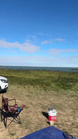 The video shows an outdoor scene with a body of water in the background. In the foreground, there is a brown and red camping chair, a white car on the left side, and a red and white bucket with a silver lid on the ground. The sky is blue with some white clouds. The landscape consists of dry grass and tall green reeds. The camera pans slightly to the right.