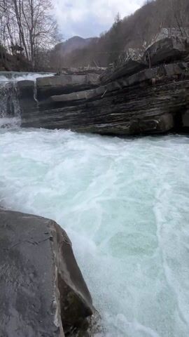 The video shows a cascading waterfall flowing over layered rock formations. The water is white and foamy as it crashes against the rocks. Bare trees line the banks of the stream. A small structure is visible in the distance. The sky is partly cloudy, with patches of blue.