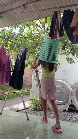 A girl wearing a green t-shirt and pink shorts, with red flip-flops, walks near a clothesline in an outdoor setting. The girl has a green plastic laundry basket on her head. The clothesline is on a metal rack and is hanging with multiple items of clothing. The backdrop includes a tin roof, a small tree, and a wall. There are also large concrete rings on the ground. The camera remains still throughout the video.