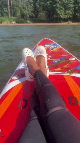 The video shows a first-person perspective of someone relaxing on a paddleboard in a body of water. The person is wearing white Crocs with decorative charms, black leggings, and has their legs crossed. The paddleboard is red and white with a colorful design. The background features a lake or river with trees and a shoreline visible in the distance. The water appears calm and the sky is clear and blue. The camera is positioned to capture the feet and legs of the person, with the paddleboard and the natural environment filling the rest of the frame. There is no visible action other than the stillness of the person on the paddleboard, suggesting a moment of relaxation or rest.