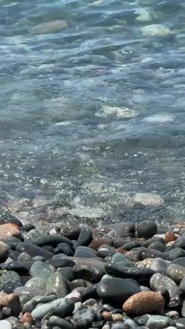The video shows a close-up view of waves washing over a rocky shore. The rocks are varied in size and color, appearing wet and smooth. The water is clear, with sunlight reflecting off its surface. The waves are gentle, creating a peaceful and natural scene. There are no people or animals visible. The environment is a beach.