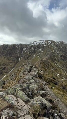 The video shows a rocky mountain path leading towards a peak partially covered in snow. The sky is overcast with a mix of dark and light clouds. To the right of the path, a mountain slope descends towards a dark, still lake. There is sparse vegetation on the slopes.